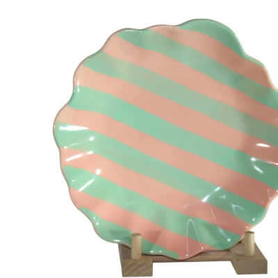 Prato de cerâmica decorativo rosa e verde água com bordos ondulados apoiado numa base de madeira.