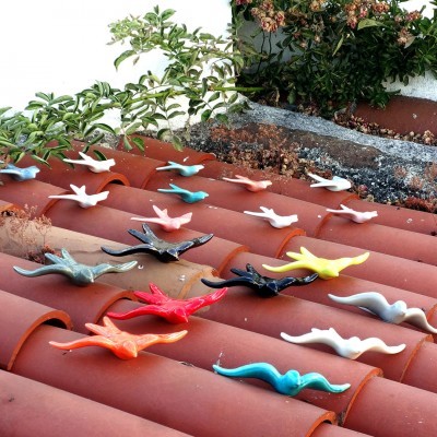 Figuras coloridas de pássaros em cima de telhas de barro vermelho com plantas ao fundo