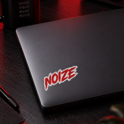 Laptop cinzento com autocolante NOIZE numa mesa preta com caneca de texto GOOD IDEAS MAKE NOISE