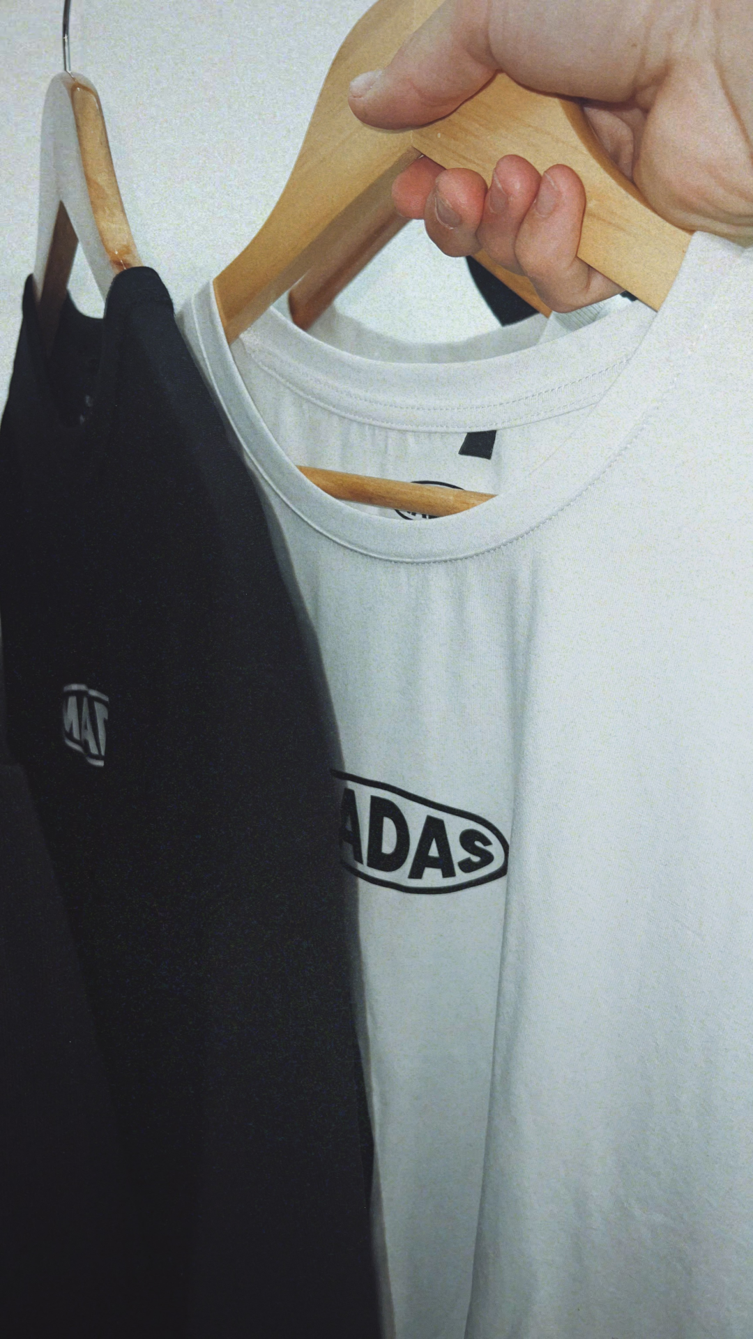 Duas t-shirts, preta e branca, penduradas em cabides de madeira numa parede clara