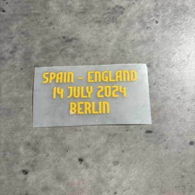 Etiqueta cinzenta com texto amarelo 'SPAIN - ENGLAND 14 JULY 2024 BERLIN' sobre superfície cinza