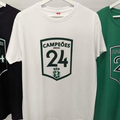 T-shirt branca com estampa verde de 'CAMPEÕES 2023/24 24 SCP' pendurada em cabide branco