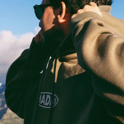 Homem de perfil com hoodie castanho e logotipo 'MADNESS' em cenário natural