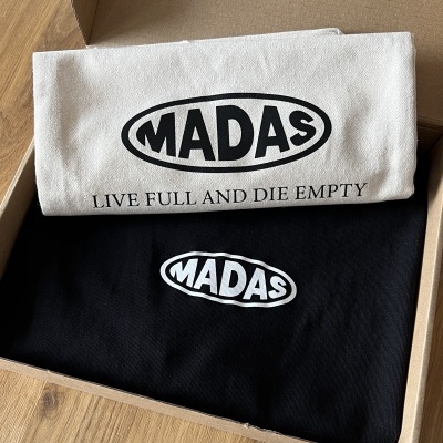 Duas t-shirts MADAS, uma branca com texto e outra preta só com logo, dentro de caixa de cartão