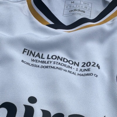 Camisola branca de futebol com gola preta e dourada e texto preto sobre a final London 2024.