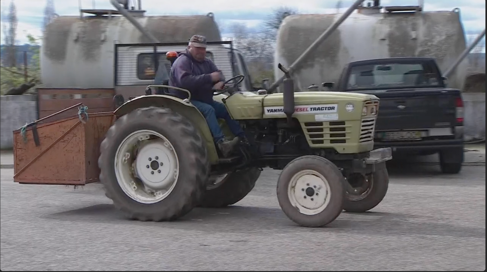 Trator verde claro com pneus grandes e pequenos em pavimento com tanque metálico e viatura ao fundo