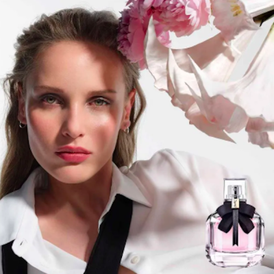 Perfume em frasco rosa claro com laço preto ao lado de mulher com camisa branca e gravata preta