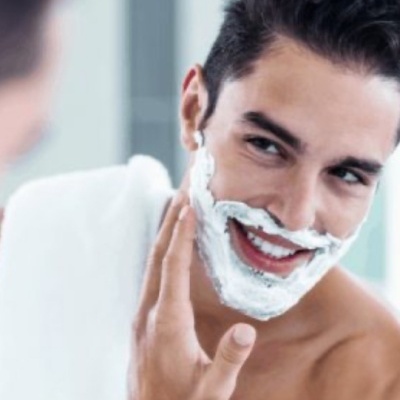 Homem a aplicar espuma de barbear branca no rosto, sorridente, frente a um espelho
