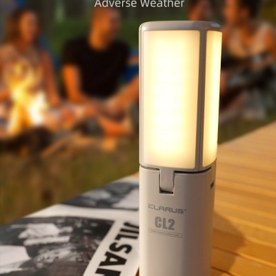 Lanterna portátil clara acesa com texto KLARUS CL2 em mesa de madeira com pessoas ao fundo