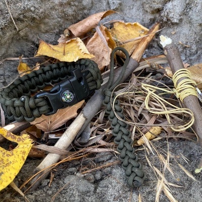 Pulseira de sobrevivência verde-oliva com bússola e pulseira trançada com cabos, sobre folhas secas e terra