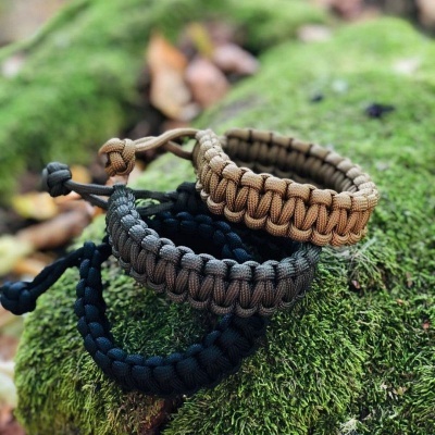 Três pulseiras trançadas de paracord em preto, castanho e bege sobre musgo verde