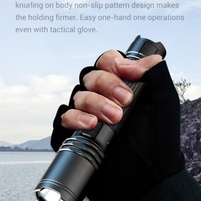 Mão com luva preta segurando lanterna táctica preta com fundo de praia e rochas