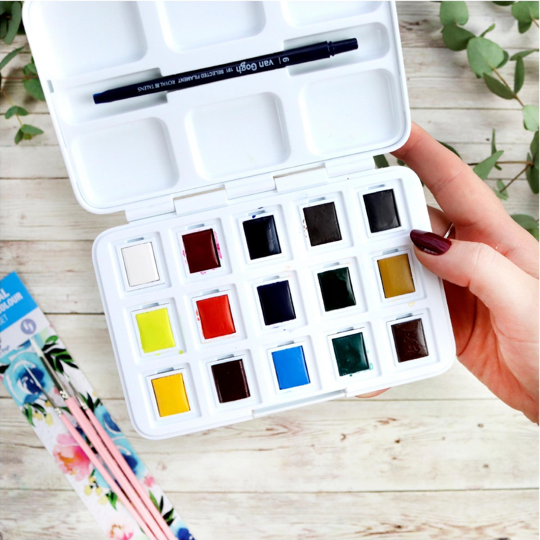 estojo de aguarelas coloridas com pincel em mão sobre mesa de madeira clara