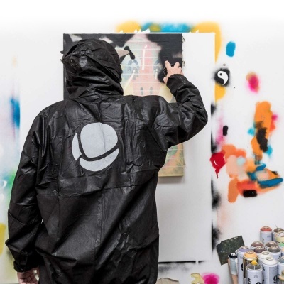 Pessoa de costas com capa preta e logotipo branco, frente a parede manchada colorida e latas de spray no chão