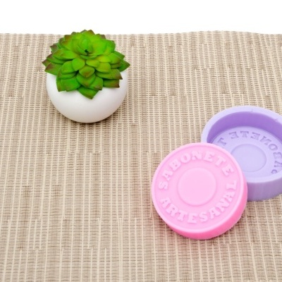 vaso branco com suculenta verde ao lado de caixa roxa com tampa rosa com texto 'SABONETE ARTESANAL' sobre tecido bege