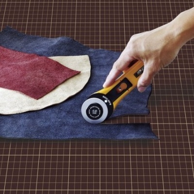 Mão com cortador circular sobre tecidos vermelho, bege e azul escuro numa mesa quadriculada.