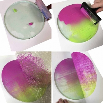 Disco redondo sendo pintado com tintas rosa e verde, espalhadas com um rodo sobre superfície branca.