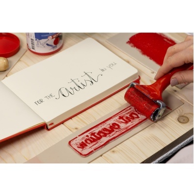 Rolo de pintura carimbando texto vermelho numa tira branca ao lado de caderno com caligrafia