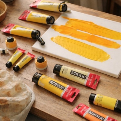 Tubos de tinta acrílica amarelos e laranjas em mesa de madeira com tela pintada e planta ao fundo.