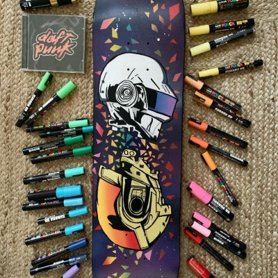 Prancha de skate com estampa de capacete e corpo robótico, rodeada por marcadores coloridos e CD Daft Punk