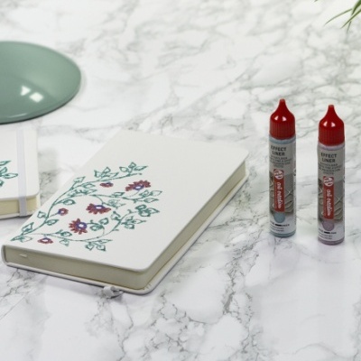 Caderno com capa decorada, tubos de tinta decorativa e prato verde sobre mesa de mármore