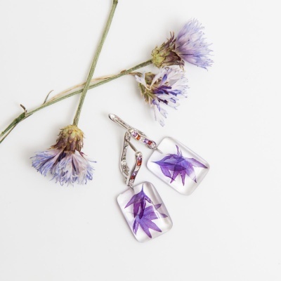 Brincos com flores roxas encapsuladas em resina e argolas prateadas com cristais ao lado de flores secas roxas
