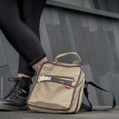 Mochila bege quadrada com bordas castanhas e bolso frontal, ao lado de pessoa com calças pretas e botas pretas, parede cinza ao fundo.