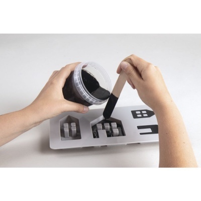 Mãos usando espátula para espalhar pó preto em formas de silicone transparentes com desenhos.