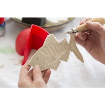 Peça de cerâmica em forma de peixe sendo gravada por uma pessoa com ferramenta de madeira