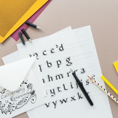 Folhas com letras caligráficas, canetas tinteiro, lápis e envelope com texto sobre fundo colorido.