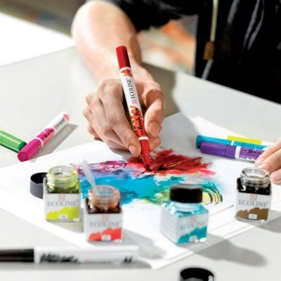 Frascos de tinta colorida e marcadores em mesa branca com mão a pintar em papel.