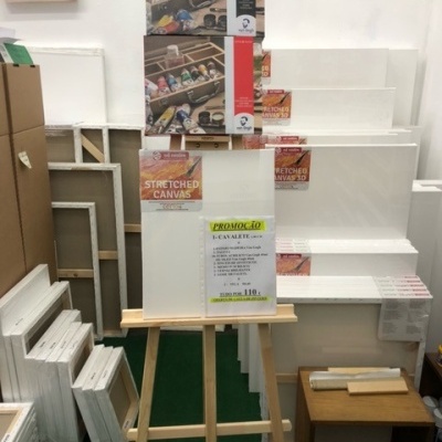 Cavalete de madeira com tela branca, etiquetas de promoção e telas empilhadas ao fundo