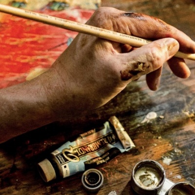 Mão com tinta a segurar pincel e tubos de tinta artística em mesa de madeira