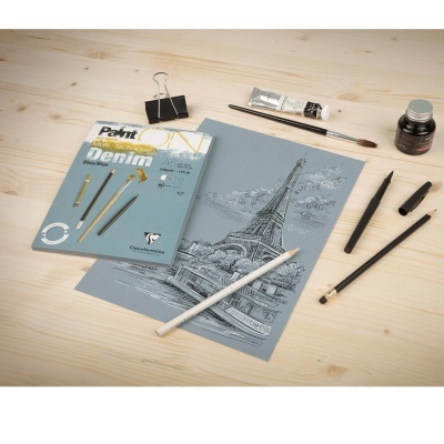 Kit de pintura com pincéis, tinta, lápis branco, tinteiro, caneta e grampo sobre mesa de madeira clara com desenho da Torre Eiffel