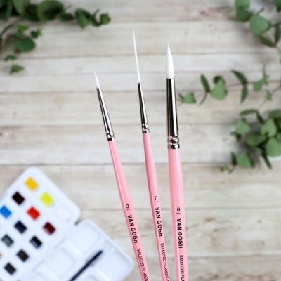 Três pincéis de pintura rosas com cerdas brancas e paleta de tintas coloridas, sobre superfície de madeira clara.
