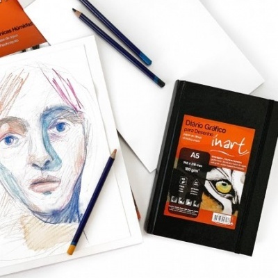 Caderno de desenho Dinart A5 com capa preta e etiqueta laranja junto a lápis azuis e desenho a lápis.