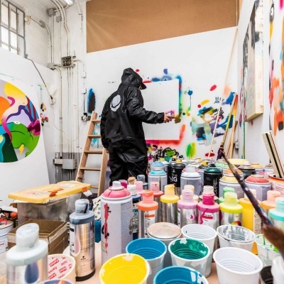 Mesa com latas de tinta spray coloridas e copos com tinta, artista de roupa preta pintando numa tela num estúdio