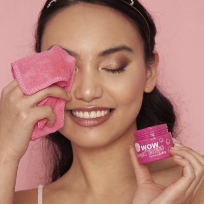 Mulher com tiara prateada a limpar o rosto com um pano rosa e segurando pote rosa de creme facial WOW