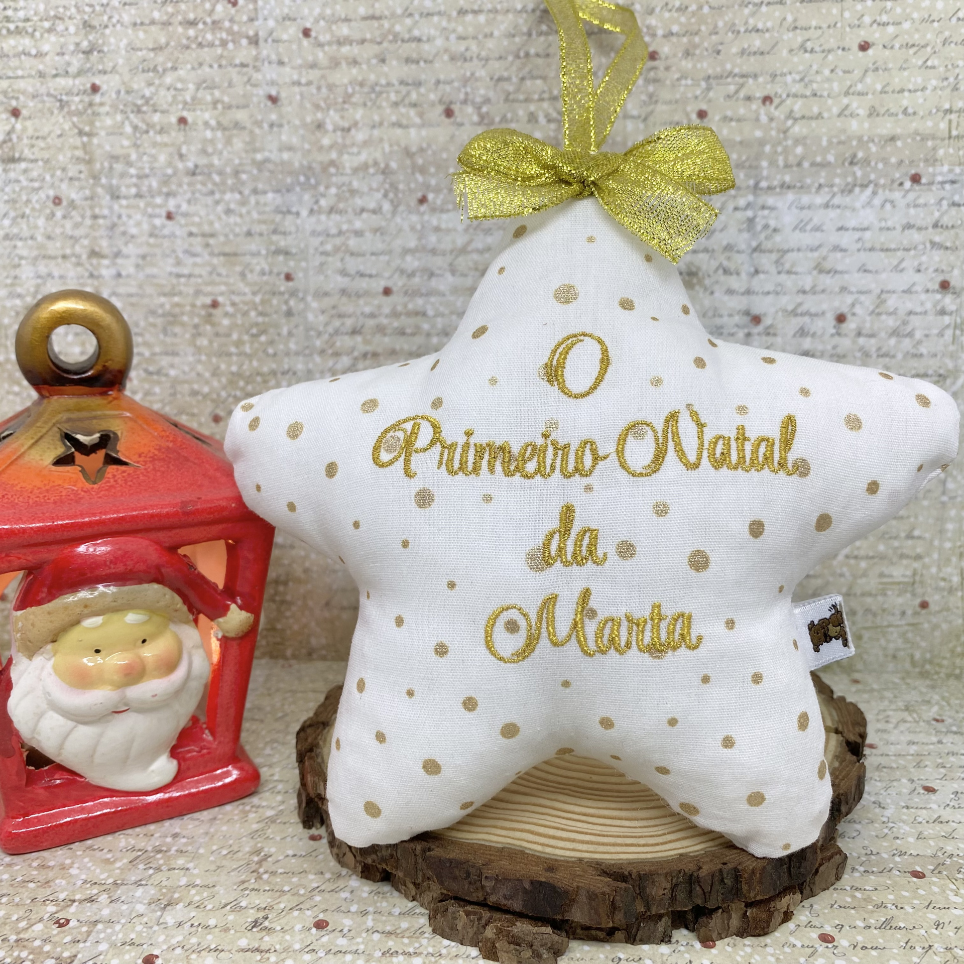 Estrela branca decorativa com pontos dourados e laço dourado, bordada com texto, junto a figurina de Pai Natal vermelha sobre fundo com padrão de texto e pontos.