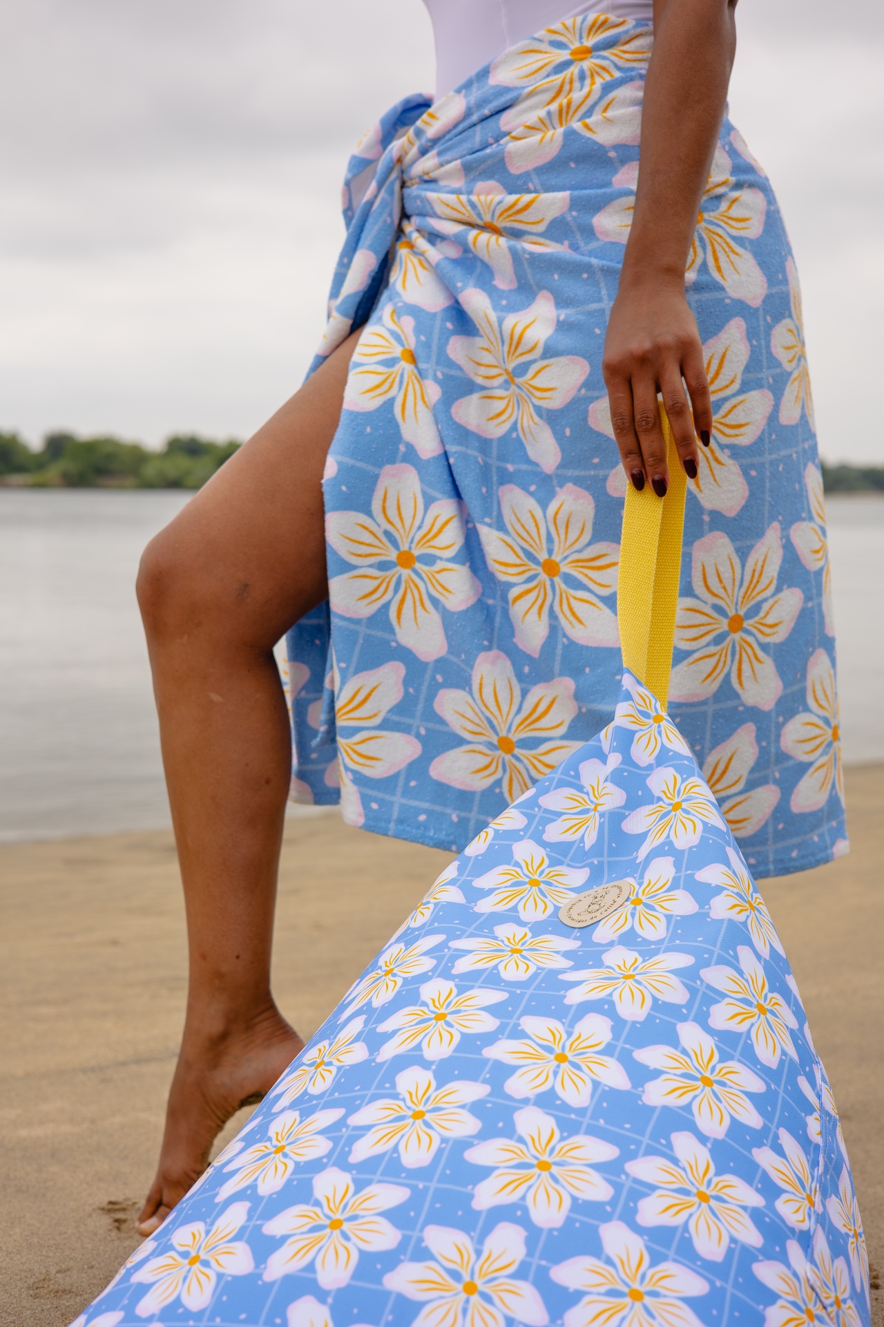 Toalha de praia azul com padrão floral amarrada na cintura e saco com padrão floral e alça amarela na praia