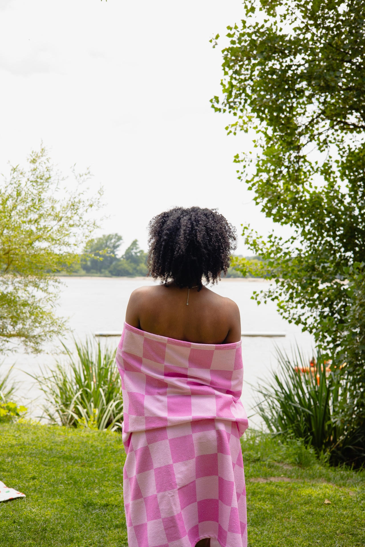 Pessoa de costas com toalha rosa xadrez ao ar livre junto a um lago e árvores