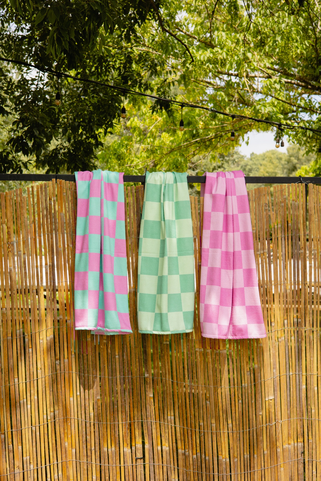 Três toalhas quadriculadas rosa e verde penduradas numa cerca de bambu com árvores ao fundo