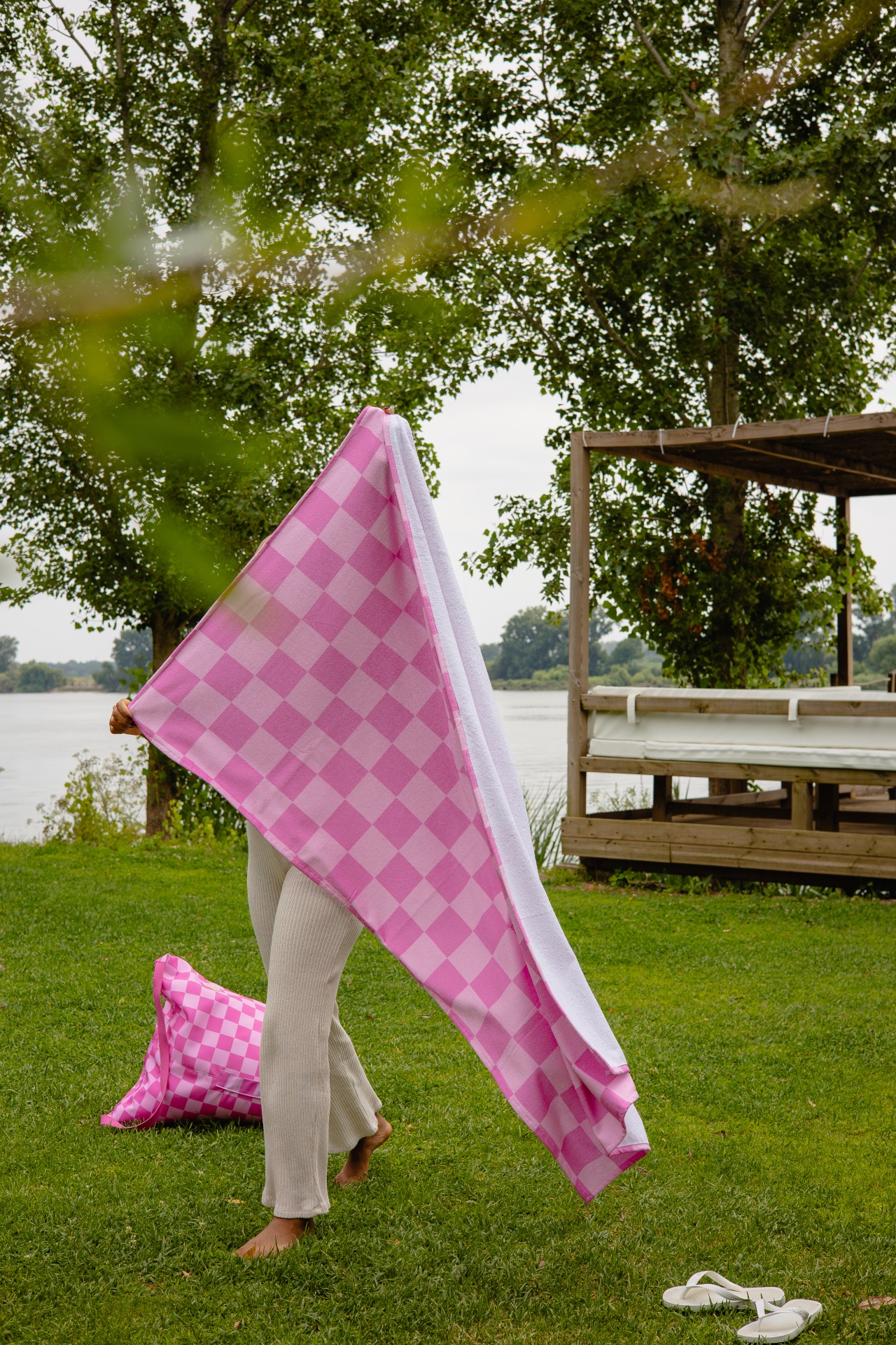 Pessoa segurando uma toalha rosa quadriculada ao ar livre numa área verde junto a um lago