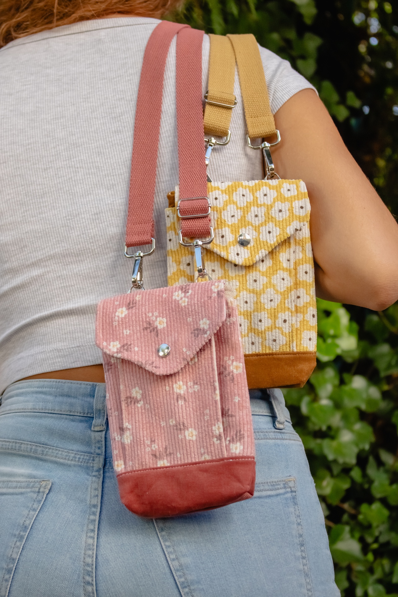 Duas bolsas pequenas de veludo cotelê com padrão floral, uma rosa e outra amarela, com alças largas, feitas em tecido ao ombro de uma pessoa.