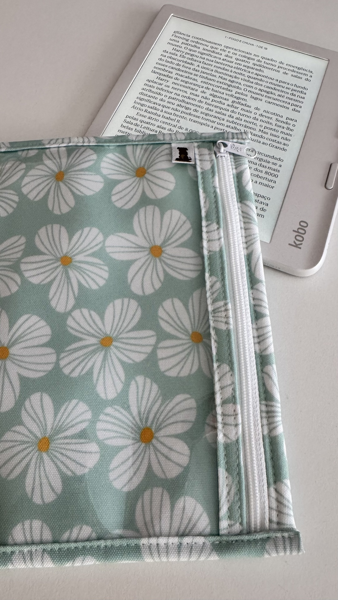 Necessaire azul claro com flores brancas e fecho branco sobre leitor Kobo com texto em português