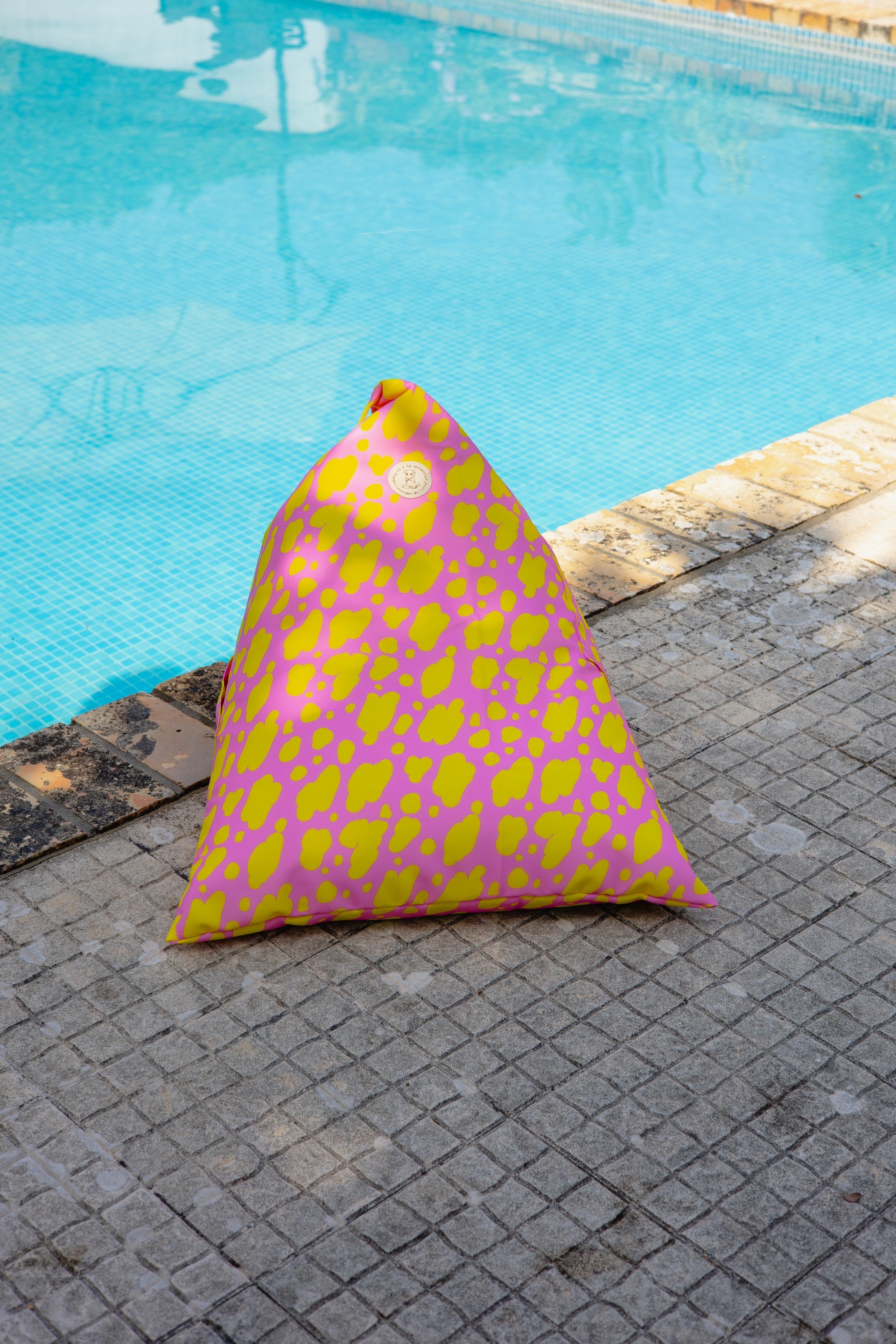 Pufe triangular cor-de-rosa com manchas amarelas junto a piscina azul