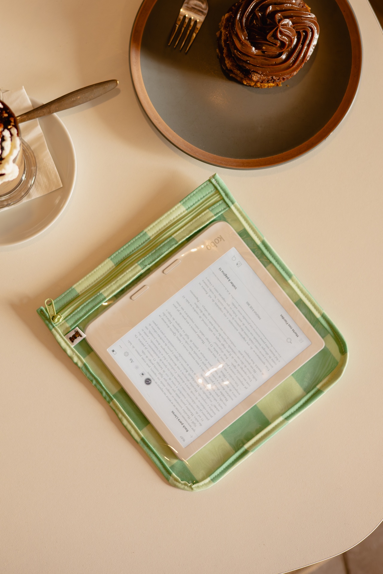 Tablet Kobo em estojo verde junto a cupcake de chocolate em prato cinza com borda castanha em mesa bege