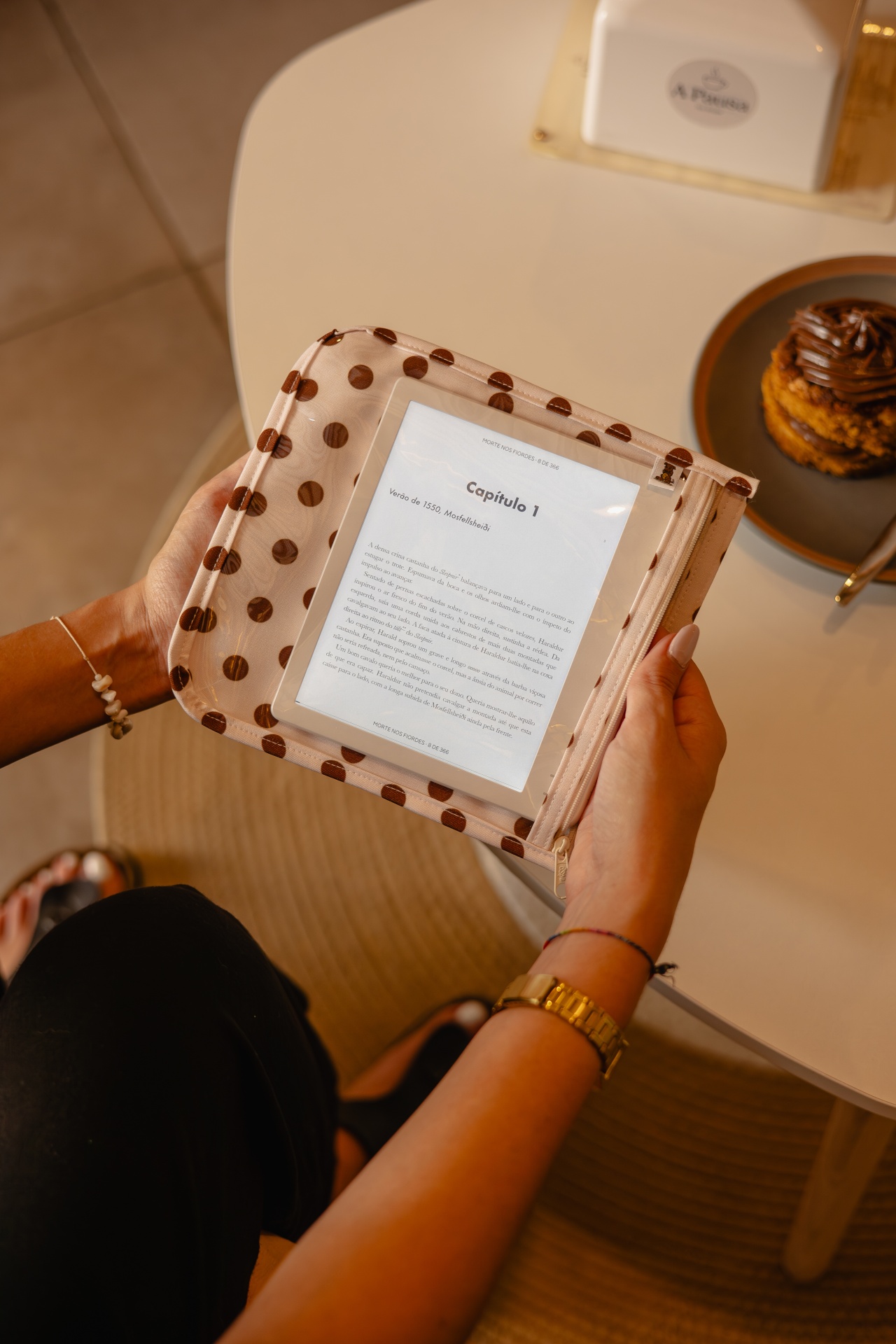 Capa para leitor digital bege com bolinhas castanhas, e-reader dentro a mostrar texto, mãos a segurá-lo junto a uma mesa com bolo e caixa de lenços.