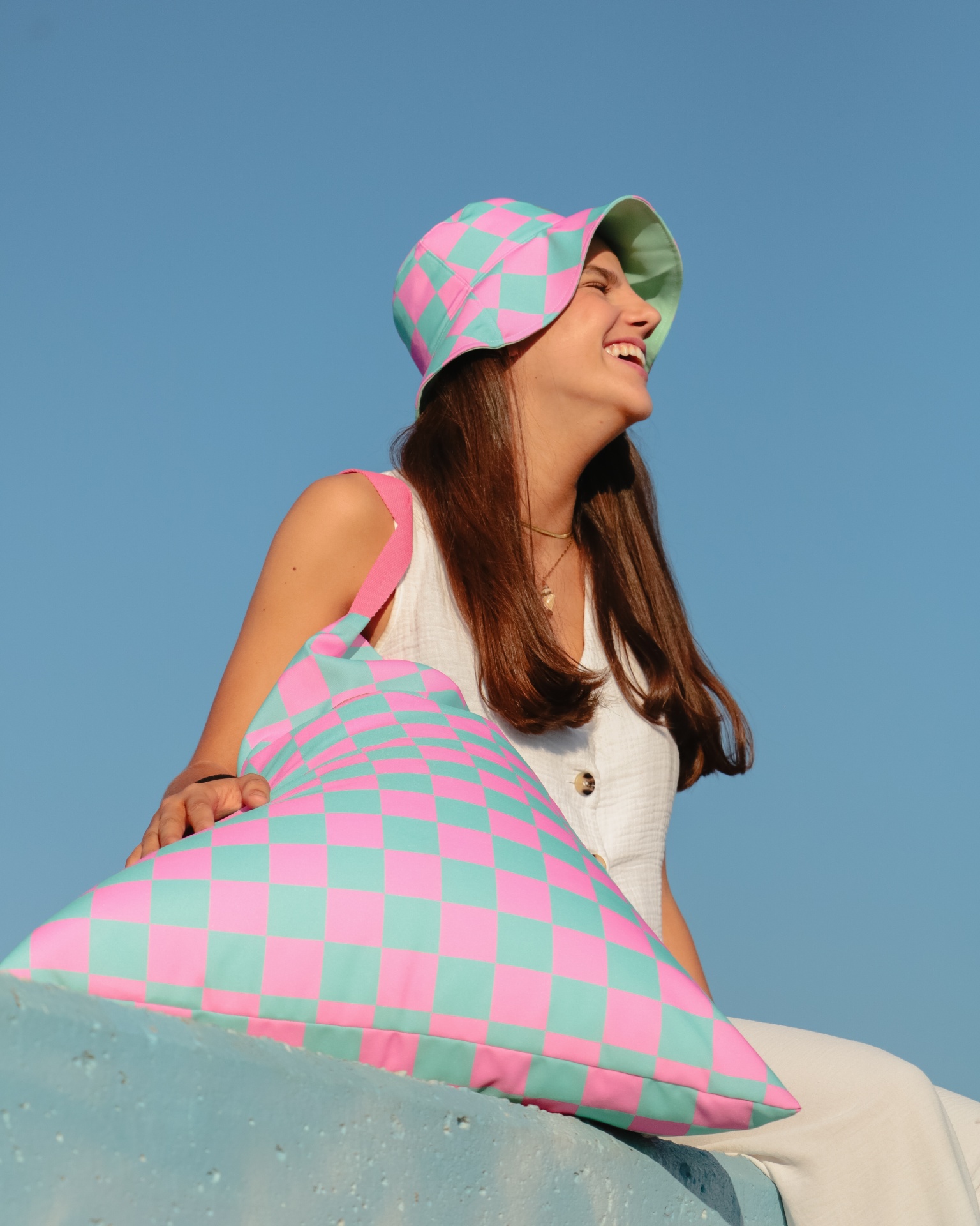 Mulher sorridente a usar chapéu de balde com padrão quadriculado em rosa e verde, segurando almofada com o mesmo padrão, céu azul de fundo.