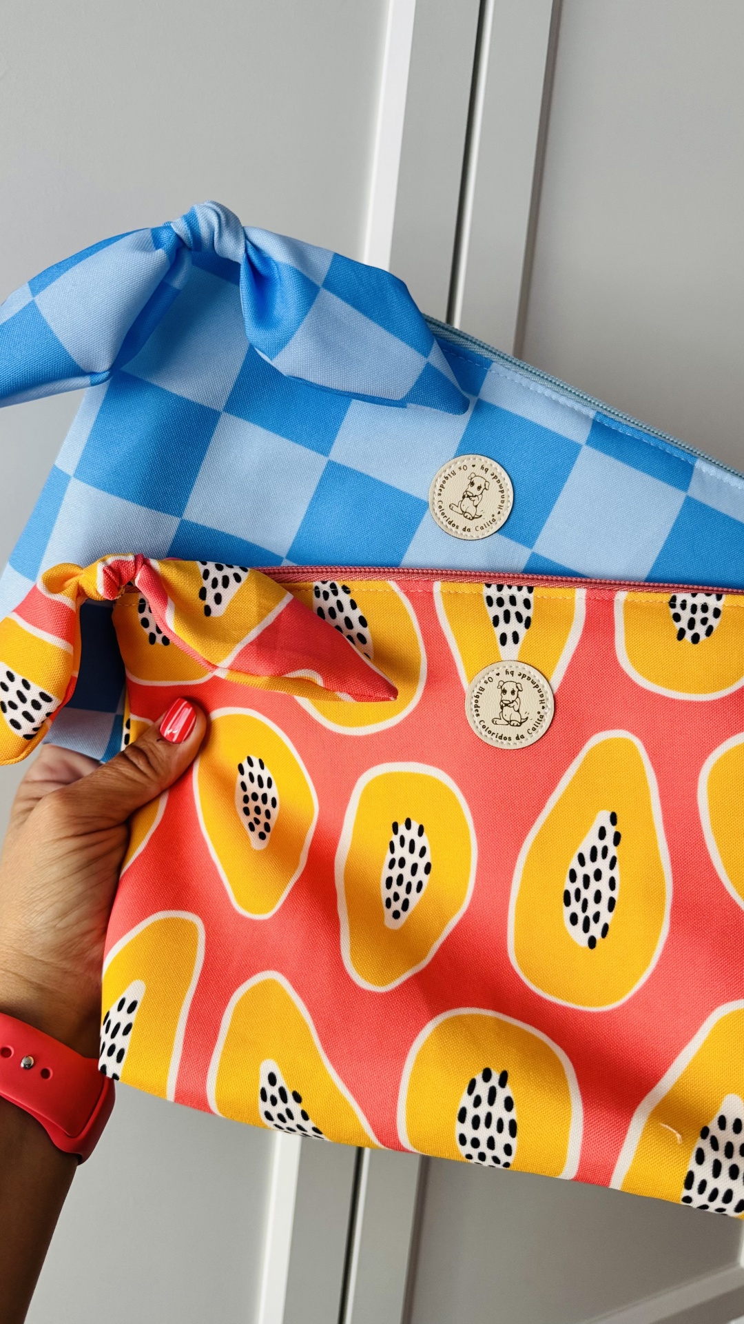 Duas bolsas de pano com padrão xadrez azul e padrão de frutas rosas, mão segurando bolsa rosa com padrão de frutas